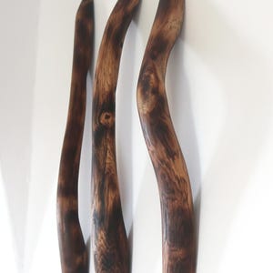Puede incluir: Tres perchas de pared de madera curvadas, hechas de madera marrón oscuro con vetas y nudos visibles. Las perchas tienen un aspecto natural y rústico y están montadas en una pared blanca. Cada percha tiene una forma orgánica única.