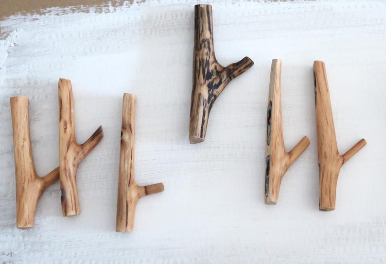 Puede incluir: Un conjunto de cinco ganchos de pared hechos de ramas de madera natural. Cada gancho presenta un acabado marr&oacute;n claro con patrones de grano de madera visibles y un peque&ntilde;o segmento de rama que sobresale. Los ganchos var&iacute;an en forma y textura, con una pieza que muestra marcas oscuras.