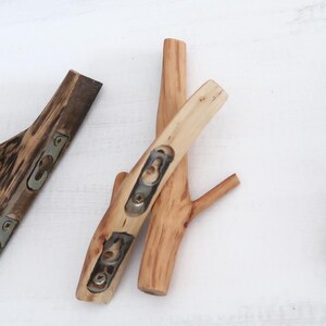 Puede incluir: Un conjunto de cuatro ganchos de pared fabricados con ramas de &aacute;rbol naturales de color marr&oacute;n claro. Cada gancho presenta una superficie de madera pulida y lisa con vetas visibles y placas de montaje met&aacute;licas integradas con ranuras.