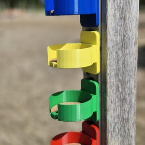 Może przedstawiać: Cztery wielokolorowe plastikowe uchwyty na kubki przymocowane do drewnianego słupa. Uchwyty są niebieskie, żółte, zielone i czerwone, z okrągłym kształtem do trzymania kubków. Uchwyty są zamontowane pionowo na słupie.