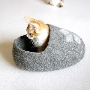 May include: A grey felt cat bed shaped like a slipper with a white paw print design. A ginger and white cat is curled up inside the bed.
