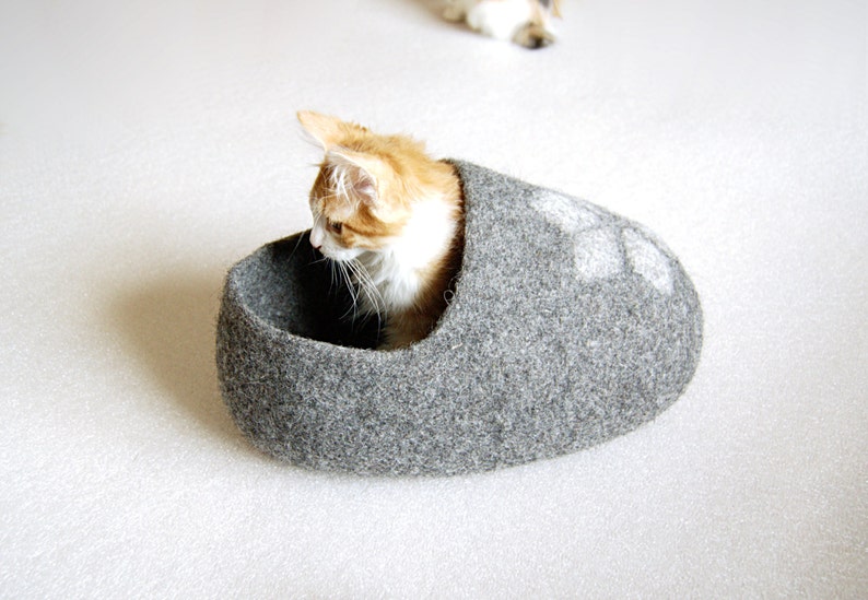 May include: A grey felt cat bed shaped like a slipper with a white paw print design. A ginger and white cat is curled up inside the bed.