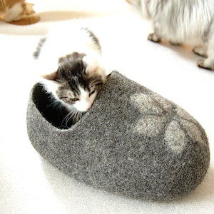May include: A grey felt cat bed shaped like a slipper with a paw print design. A white and grey kitten is curled up inside the bed.