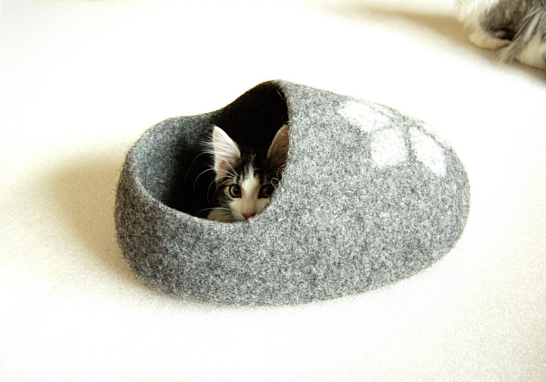 May include: A grey felt cat bed shaped like a slipper with a white paw print design. A black and white kitten is peeking out of the bed.