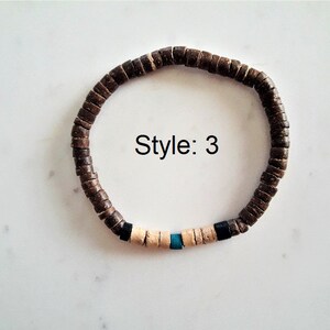 Pode incluir: Uma pulseira circular composta por pequenas contas planas em forma de disco castanho-escuro. Uma sec&ccedil;&atilde;o apresenta duas contas pretas, quatro contas bege-claras e uma conta central azul-turquesa. O texto "Style: 3" est&aacute; impresso a preto.