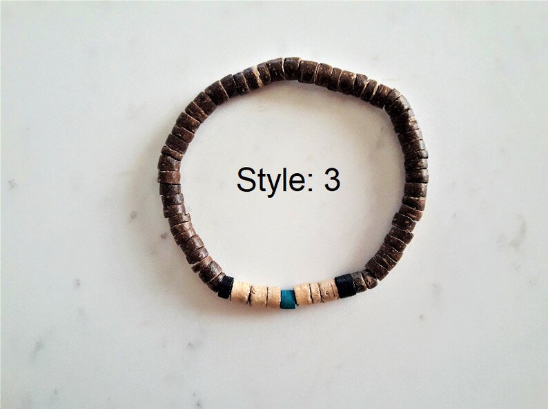 Pode incluir: Uma pulseira circular composta por pequenas contas planas em forma de disco castanho-escuro. Uma sec&ccedil;&atilde;o apresenta duas contas pretas, quatro contas bege-claras e uma conta central azul-turquesa. O texto "Style: 3" est&aacute; impresso a preto.