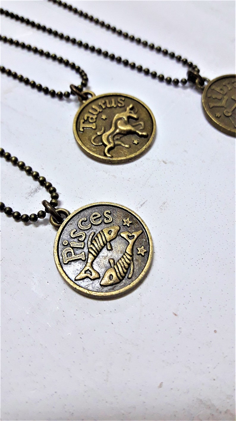 May include: Two gold-toned zodiac necklaces with black chains. The necklaces feature a circular pendant with the zodiac signs Taurus and Pisces.