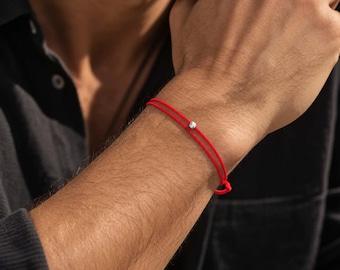 May include: A double-strand red cord bracelet featuring a single small, polished silver-toned metallic bead centered on the top. The bracelet is thin and minimalist in construction, wrapping twice around the wrist with a simple knot closure visible.