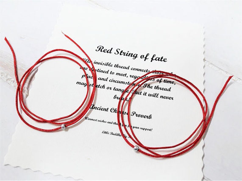 Op de afbeelding: Rood draad van het lot armband met zilveren kralen. De armband ligt op een witte achtergrond met een citaat over de rode draad van het lot. "Een onzichtbare draad verbindt degenen die bestemd zijn om elkaar te ontmoeten, ongeacht tijd, plaats en omstandigheden. De draad kan uitrekken of verstrengelen, maar hij zal nooit breken. Oud Chinees spreekwoord."