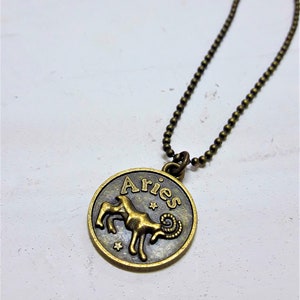 May include: A gold-toned necklace with a round pendant featuring the zodiac sign Aries. The pendant has the word "Aries" written on it in a cursive font.