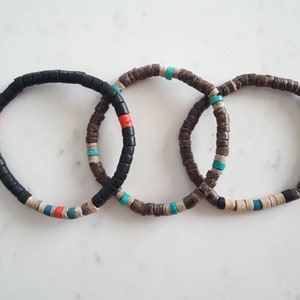 Pode incluir: Tr&ecirc;s pulseiras circulares feitas de pequenas contas planas em forma de disco. A primeira &eacute; principalmente preta com detalhes em vermelho, turquesa e bege. A segunda e a terceira s&atilde;o castanhas com contas turquesa e bege intercaladas.