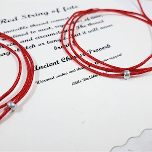 Op de afbeelding: Rode touw armbanden met zilveren kralen. De armbanden zijn aan elkaar gebonden en liggen op een witte achtergrond met tekst die "Red String of Fate" en "Ancient Chinese Proverb" zegt.