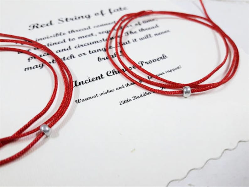 Op de afbeelding: Rode touw armbanden met zilveren kralen. De armbanden zijn aan elkaar gebonden en liggen op een witte achtergrond met tekst die "Red String of Fate" en "Ancient Chinese Proverb" zegt.