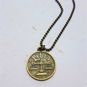 May include: A gold-toned necklace with a round pendant featuring the zodiac symbol for Libra and the word "Libra" inscribed on it.