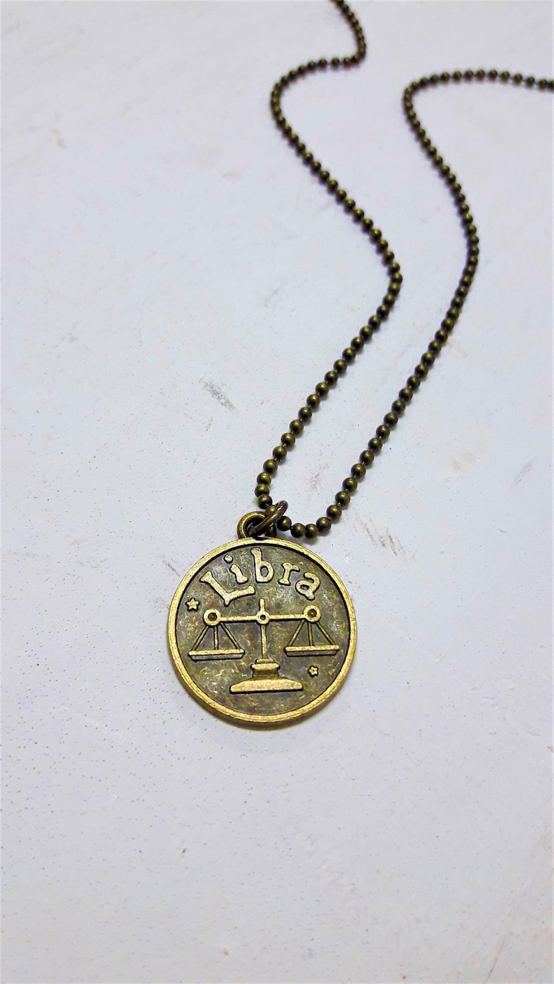May include: A gold-toned necklace with a round pendant featuring the zodiac symbol for Libra and the word "Libra" inscribed on it.