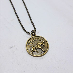 May include: A gold-toned necklace with a round pendant featuring the zodiac sign Taurus. The pendant has the word "Taurus" written around the edge and a depiction of a bull inside.