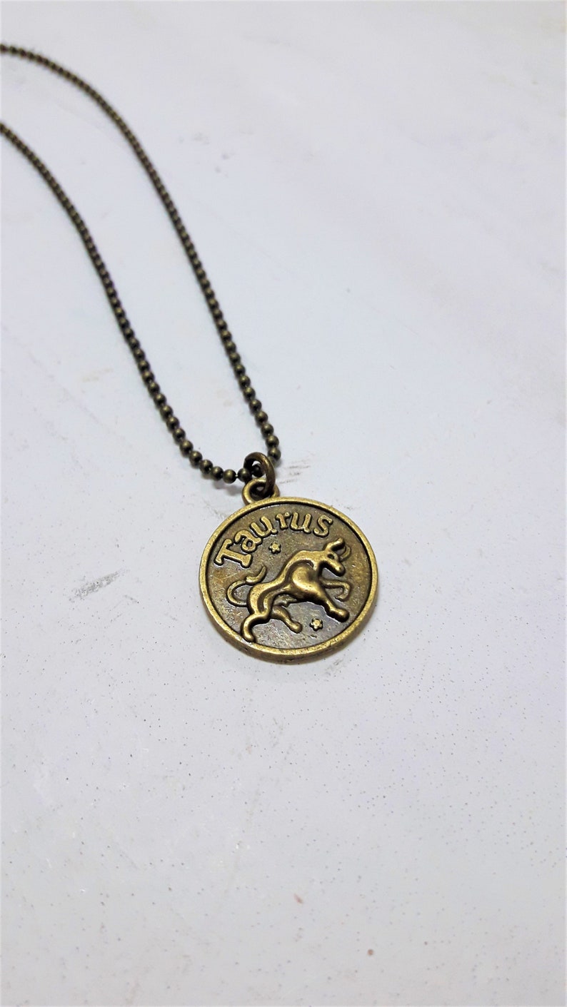 May include: A gold-toned necklace with a round pendant featuring the zodiac sign Taurus. The pendant has the word "Taurus" written around the edge and a depiction of a bull inside.