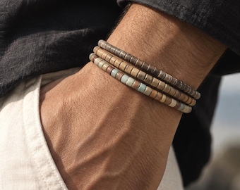 Pode incluir: Um conjunto de três pulseiras empilhadas com pequenas contas cilíndricas em tons terrosos de castanho, bege e azul-esverdeado suave. As contas estão dispostas num padrão uniforme, criando um visual em camadas no pulso com textura.