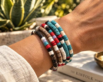 Pode incluir: Um conjunto de cinco pulseiras feitas de pequenas contas cilíndricas em várias cores, incluindo turquesa, branco, vermelho, preto, castanho e roxo claro. As pulseiras são usadas juntas no pulso, apresentando um padrão repetitivo destas contas coloridas dispostas num design circular de vários fios.