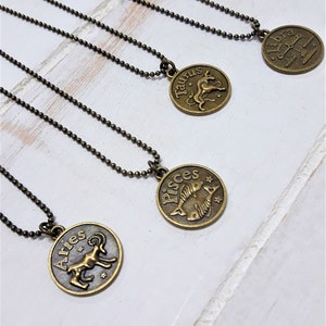 May include: Four gold-toned necklaces with zodiac signs pendants. The pendants are Aries, Taurus, Pisces, and Libra. The necklaces are on a white wooden surface.
