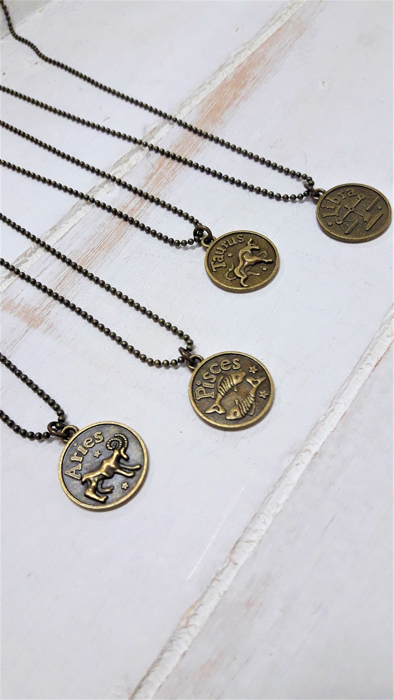 May include: Four gold-toned necklaces with zodiac signs pendants. The pendants are Aries, Taurus, Pisces, and Libra. The necklaces are on a white wooden surface.