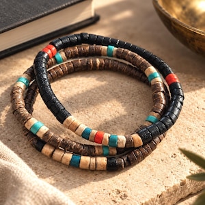 Pode incluir: Um conjunto de tr&ecirc;s pulseiras empilhadas feitas de pequenas contas cil&iacute;ndricas em forma de disco. As contas s&atilde;o coloridas em preto, castanho escuro, bege claro, turquesa brilhante e laranja vibrante, dispostas em padr&otilde;es alternados ao longo de cada fio.