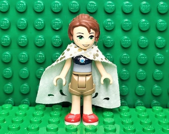 May include: A Lego minifigure of a girl with brown hair, green eyes, and a white cape with gold accents. She is wearing a blue and white patterned shirt, brown shorts, and red shoes.