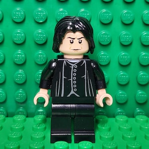 May include: A Lego minifigure of a character with black hair and a stern expression. The figure wears a black jacket with a silver buttoned design over a white shirt, and black pants. The figure stands on a green Lego base.