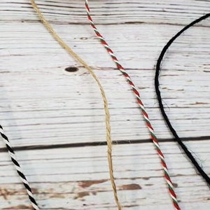 May include: Four different types of twine, including black and white, red and white, natural, and black. The twine is laid out on a white wooden surface.