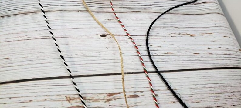 May include: Four different types of twine, including black and white, red and white, natural, and black. The twine is laid out on a white wooden surface.