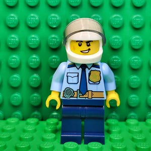 May include: A Lego police officer minifigure with a light blue shirt, dark blue pants, and a silver helmet. The figure has a yellow face with a smile and is standing on a green Lego baseplate.