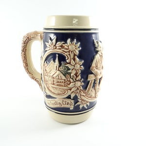 May include: A ceramic beer stein with a blue and white design. The stein features a scene of a church and a man with a pickaxe. The text "ein lustig Lied" is written on the stein.