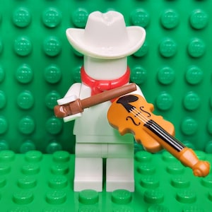 May include: A white Lego minifigure wearing a white cowboy hat and a red scarf, holding a brown violin and a brown bow.