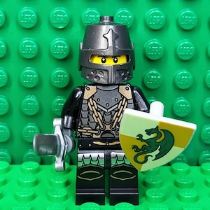 May include: A Lego knight minifigure with a silver helmet, armor, and a shield featuring a green dragon. The knight holds a silver sword and stands on a green Lego baseplate. The figure has yellow eyes.