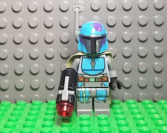 May include: A blue and gray Lego minifigure of a Mandalorian with a blaster rifle. The minifigure has a blue helmet with a purple splatter design and a green cape.