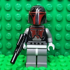 May include: A Lego minifigure of a character with a black and red helmet and armor, holding a black blaster rifle. The figure has gray legs and arms, and white hands. The background is a green Lego baseplate.