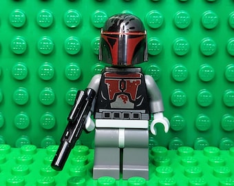 May include: A Lego minifigure of a character with a black and red helmet and armor, holding a black blaster rifle. The figure has gray legs and arms, and white hands. The background is a green Lego baseplate.