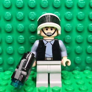 May include: A Lego minifigure of a character wearing a white helmet with a black visor, a black vest over a light blue shirt, and gray pants. The figure is holding a black and gray blaster with a blue tip. The background is a green Lego baseplate.