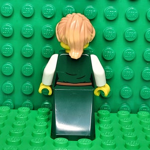 May include: A Lego minifigure with blonde hair, wearing a white shirt and a long green dress with a brown belt.
