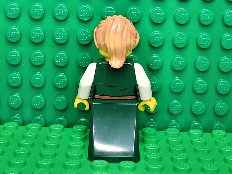 May include: A Lego minifigure with blonde hair, wearing a white shirt and a long green dress with a brown belt.