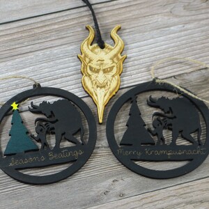 May include: Two black wooden Christmas ornaments with a silhouette of a Krampus figure and a child. The ornaments are round and have a string for hanging. The text on the ornaments reads "Seasons Greetings" and "Merry Krampusnacht".