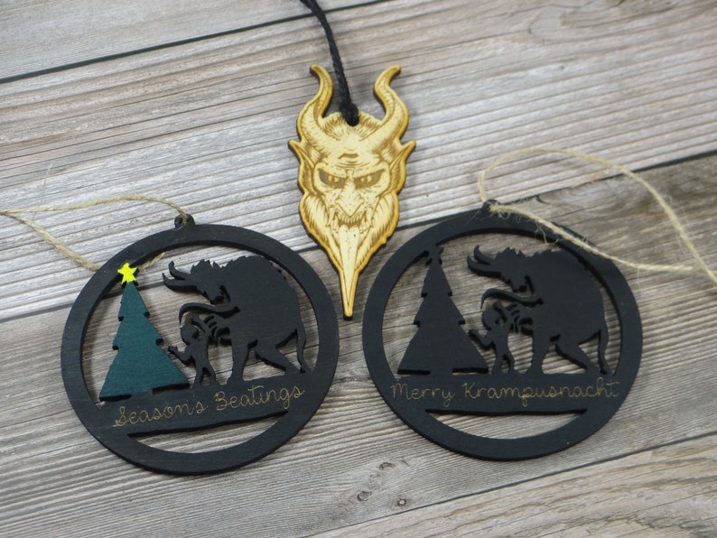 May include: Two black wooden Christmas ornaments with a silhouette of a Krampus figure and a child. The ornaments are round and have a string for hanging. The text on the ornaments reads "Seasons Greetings" and "Merry Krampusnacht".