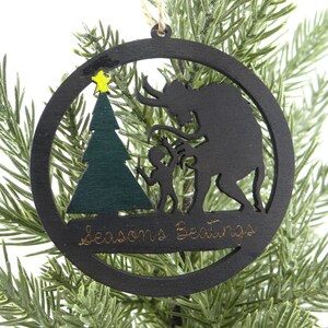 May include: A black wooden Christmas ornament with a silhouette of a monster, a child, and a Christmas tree. The ornament has the text "Seasons Greetings" written on it.