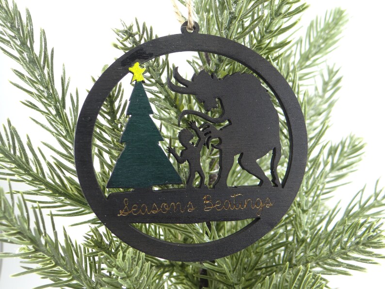 May include: A black wooden Christmas ornament with a silhouette of a monster, a child, and a Christmas tree. The ornament has the text "Seasons Greetings" written on it.