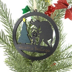 May include: A black wooden Christmas ornament with a silhouette of a monster, a tree, and a child. The ornament is hanging on a green artificial Christmas tree. The ornament has the text "Season's Greetings" on it.