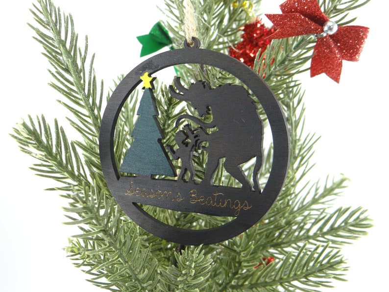 May include: A black wooden Christmas ornament with a silhouette of a monster, a tree, and a child. The ornament is hanging on a green artificial Christmas tree. The ornament has the text "Season's Greetings" on it.