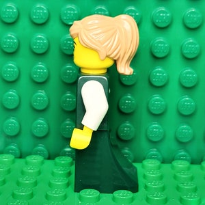 May include: A Lego minifigure of a woman with brown hair in a ponytail, wearing a white shirt and a dark green skirt.