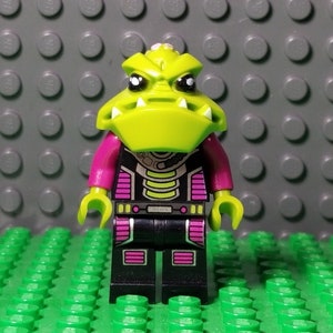 May include: A Lego minifigure of an alien creature with a lime green head and a stern expression. The figure has magenta arms, black legs, and a black torso with magenta and yellow accents. The figure is standing on a green Lego base.