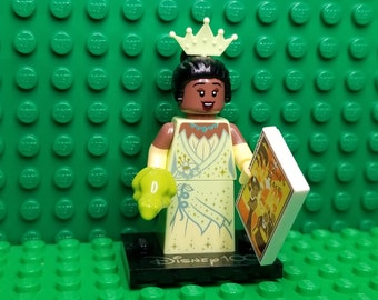 May include: A LEGO minifigure of Princess Tiana from Disney's The Princess and the Frog. She is wearing a yellow and green dress with a crown and holding a green frog and a book that says "Tiana's Place".