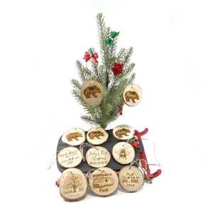 May include: A set of 9 wood slice ornaments with various designs, including a bear, a bee, a tree, a house, and text. The ornaments are hanging on a small artificial Christmas tree and a red sled.